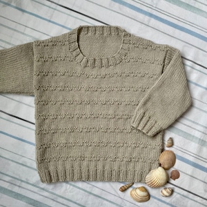 May include: A beige knitted sweater with a round neckline. The sweater is laid flat on a striped fabric background.