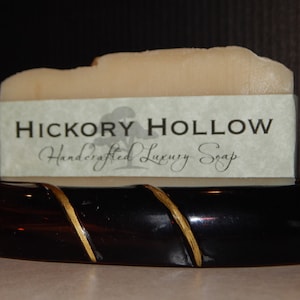 Puede incluir: Una pastilla de jabón beige claro con una etiqueta que dice "Hickory Hollow Handcrafted Luxury Soap". Descansa sobre un jabonera marrón oscuro con un acento dorado. El jabón mide unos 10 cm de largo.