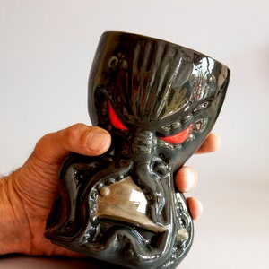 May include: A black ceramic mug shaped like an octopus with red eyes. The mug is being held in a hand.