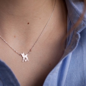 May include: A silver-toned necklace featuring a small, flat pendant shaped like a standing fawn. The pendant is attached to a delicate, thin silver-coloured chain that rests against the collarbone, creating a simple and clean aesthetic.