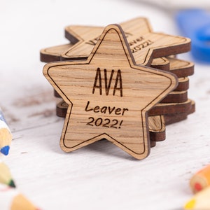 May include: A stack of wooden star-shaped keepsakes. Each star is engraved with the name "AVA" and the text "Leaver 2022!". The stars are made of light brown wood with a darker brown edge.
