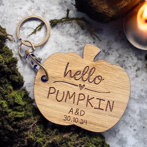 Peut inclure: Porte-clés en bois en forme de citrouille avec le texte "hello pumpkin" et un petit cœur. Le porte-clés comprend également les initiales "A&D" et la date "30.10.24".