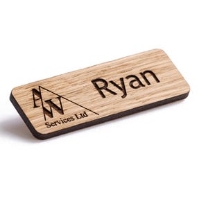 以下が含まれることがあります： 黒い縁取りのある木製のネームタグ。ネームタグには、「A」と「W」の文字と「Services Ltd」というテキストのロゴがあります。右側に「Ryan」という名前が刻まれています。