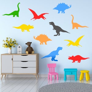 May include: A light blue wall with colorful dinosaur silhouettes. The dinosaurs are in various poses and include a pterodactyl, a stegosaurus, a triceratops, and a tyrannosaurus rex. There is a white dresser with three drawers, a pink child's chair, and a blue side table in the room.