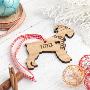 May include: Wooden Christmas ornament shaped like a dog wearing a Santa hat and scarf. The ornament is personalized with the name "Pepper".