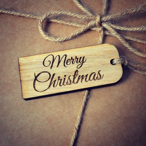 Könnte beinhalten: Ein braunes Papierpaket, das mit Schnur gebunden ist und einen hölzernen Geschenkanhänger mit der Aufschrift "Frohe Weihnachten" trägt.