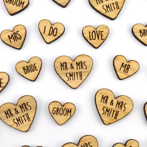 Può includere: Bomboniere a forma di cuore in legno con varie frasi incise come "I DO", "LOVE", "MR & MRS SMITH", "BRIDE" e "GROOM". I cuori hanno una venatura naturale del legno e un bordo scuro.