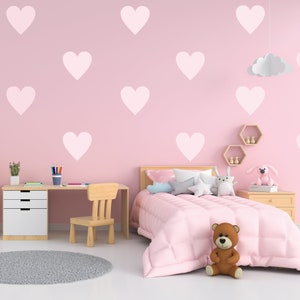 Op de afbeelding: Een roze slaapkamer met een witte wolklamp, een bed met roze beddengoed en een teddybeer, een bureau met een stoel en een roze muur met witte hart stickers.