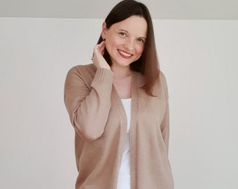 May include: A long, light brown cardigan with a V-neck and long sleeves. The cardigan is open and worn over a white tank top and white jeans.