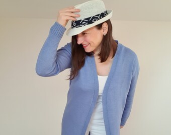 May include: A woman wearing a light blue cardigan with a white tank top and white pants. She is wearing a white fedora with a black and white patterned band.