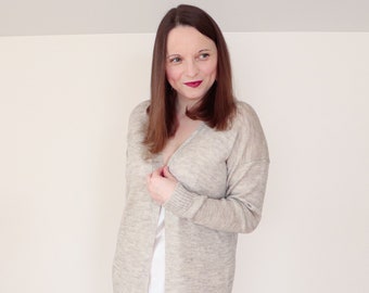 May include: A woman wearing a long, light gray cardigan with a white satin slip dress underneath. The cardigan is open and the woman is holding the front of it with her right hand.