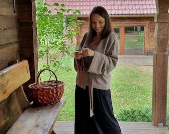 Op de afbeelding: Een vrouw staat op een houten veranda, gekleed in een beige trui en een zwarte broek. Een gevlochten mand staat op een houten bank. Een rood pannendak van een gebouw is zichtbaar op de achtergrond.