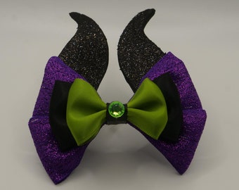May include: Purple and black glitter bow with green ribbon and a green gem in the center. The bow has two black horns on top.