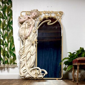 Huge Art Nouveau Mucha Style Iron Mirror with a life-size plaster resin statue of a 66” tall young woman in a flowing cream-colored dress. The arched mirror on the right side of the woman is surrounded by a wrought iron frame with vines and flowers along the top.