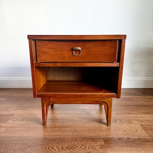 May include: A medium-brown wooden nightstand featuring a single top drawer with a centered metallic pull handle. Below the drawer is an open storage shelf. The piece is supported by four tapered legs that extend from the base of the unit.