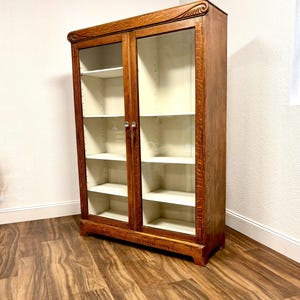 K&ouml;nnte beinhalten: Ein antiker B&uuml;cherregal aus Holz mit Glast&uuml;ren. Das B&uuml;cherregal hat eine warme braune Oberfl&auml;che, Glast&uuml;ren und mehrere wei&szlig;e Regale. Die Oberseite des B&uuml;cherregals weist ein dekoratives Schnitzmuster auf. Es ist ein klassisches M&ouml;belst&uuml;ck.