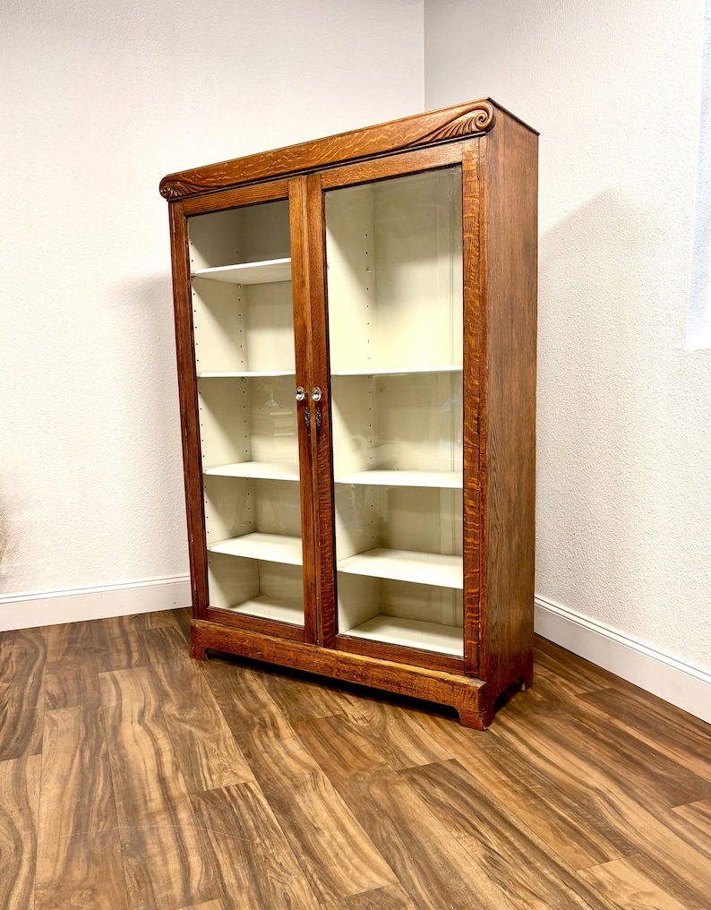K&ouml;nnte beinhalten: Ein antiker B&uuml;cherregal aus Holz mit Glast&uuml;ren. Das B&uuml;cherregal hat eine warme braune Oberfl&auml;che, Glast&uuml;ren und mehrere wei&szlig;e Regale. Die Oberseite des B&uuml;cherregals weist ein dekoratives Schnitzmuster auf. Es ist ein klassisches M&ouml;belst&uuml;ck.