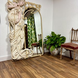 May include: A large, ornate mirror with a white, sculpted frame featuring a woman in a flowing gown. The mirror has an arched top and is surrounded by intricate floral details.