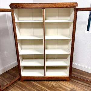 K&ouml;nnte beinhalten: Ein B&uuml;cherregal aus Holz mit Glast&uuml;ren, mit einem dunkelbraunen Rahmen und einem hellfarbigen Innenraum. Das B&uuml;cherregal hat mehrere Regale und ist offen, was seine Speicherkapazit&auml;t zeigt. Die Oberseite des B&uuml;cherregals hat ein dekoratives Schnitzmuster.