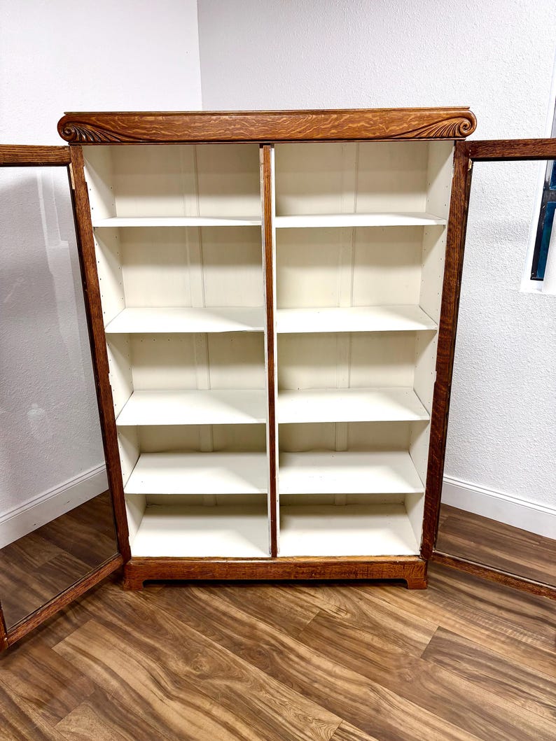 K&ouml;nnte beinhalten: Ein B&uuml;cherregal aus Holz mit Glast&uuml;ren, mit einem dunkelbraunen Rahmen und einem hellfarbigen Innenraum. Das B&uuml;cherregal hat mehrere Regale und ist offen, was seine Speicherkapazit&auml;t zeigt. Die Oberseite des B&uuml;cherregals hat ein dekoratives Schnitzmuster.