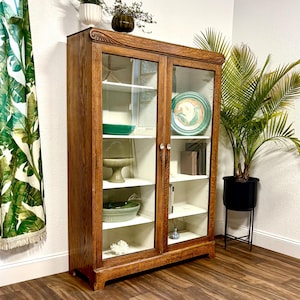 Antique Oak Cabinet with glass doors. It is brown, made of solid Oak Wood with a beautiful woodgrain. The cabinet has a white painted interior with a center divider and there are 4 adjustable white shelves on each side. It is filled with decorative items, including plates, books, and vases. A green plant in a black pot sits nearby. The cabinet is brown with white shelves and a white interior. The floor is hardwood and the walls are white. There is a green banana leaf curtain on the left.