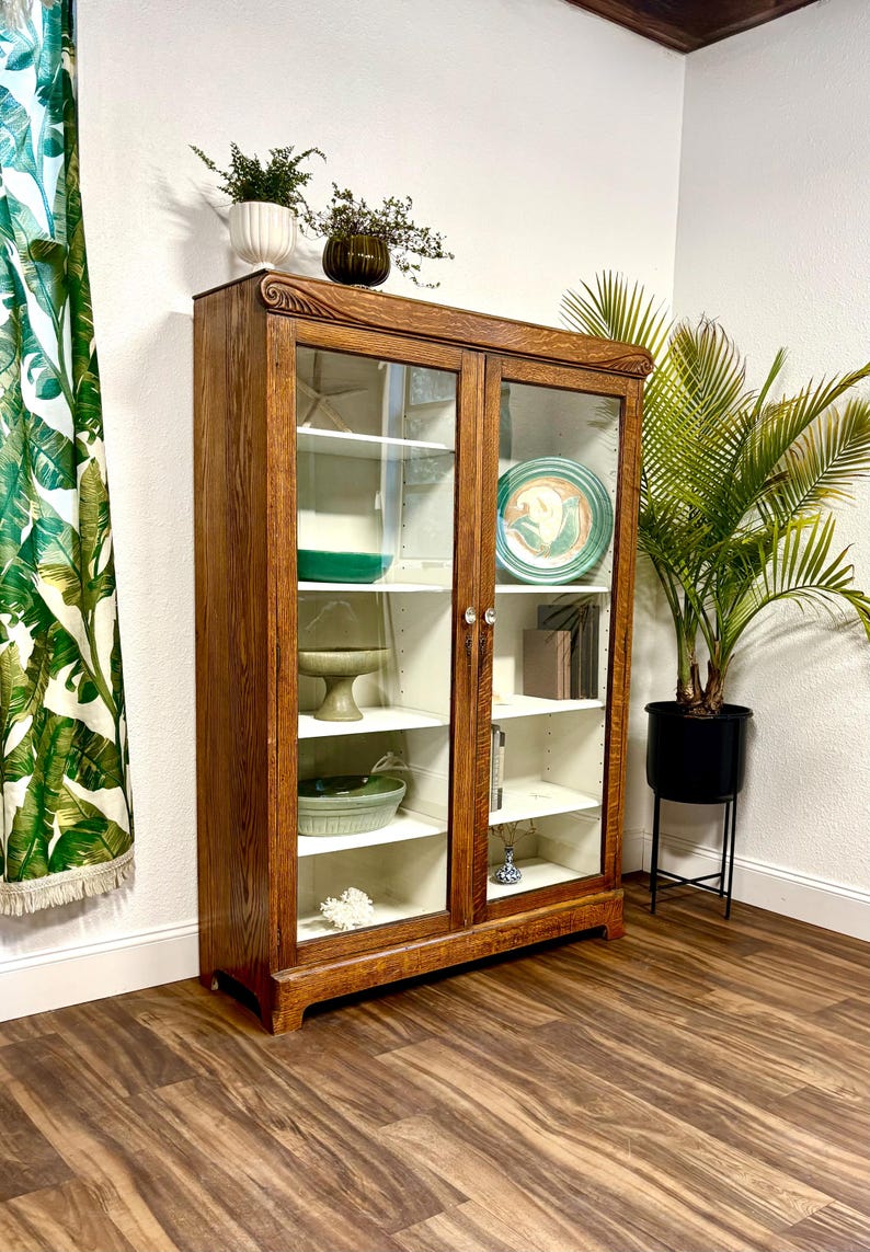 Antique Oak Cabinet with glass doors. It is brown, made of solid Oak Wood with a beautiful woodgrain. The cabinet has a white painted interior with a center divider and there are 4 adjustable white shelves on each side. It is filled with decorative items, including plates, books, and vases. A green plant in a black pot sits nearby. The cabinet is brown with white shelves and a white interior. The floor is hardwood and the walls are white. There is a green banana leaf curtain on the left.