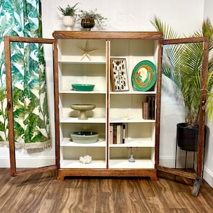 K&ouml;nnte beinhalten: Ein Vintage-Holzschrank mit Glast&uuml;ren, der verschiedene dekorative Gegenst&auml;nde pr&auml;sentiert. Der Schrank hat eine warme braune Oberfl&auml;che, wei&szlig;e Innenregale und enth&auml;lt einen Seestern, gr&uuml;ne Schalen, B&uuml;cher und eine gerahmte Insektenausstellung. Eine Topfpflanze ist sichtbar.