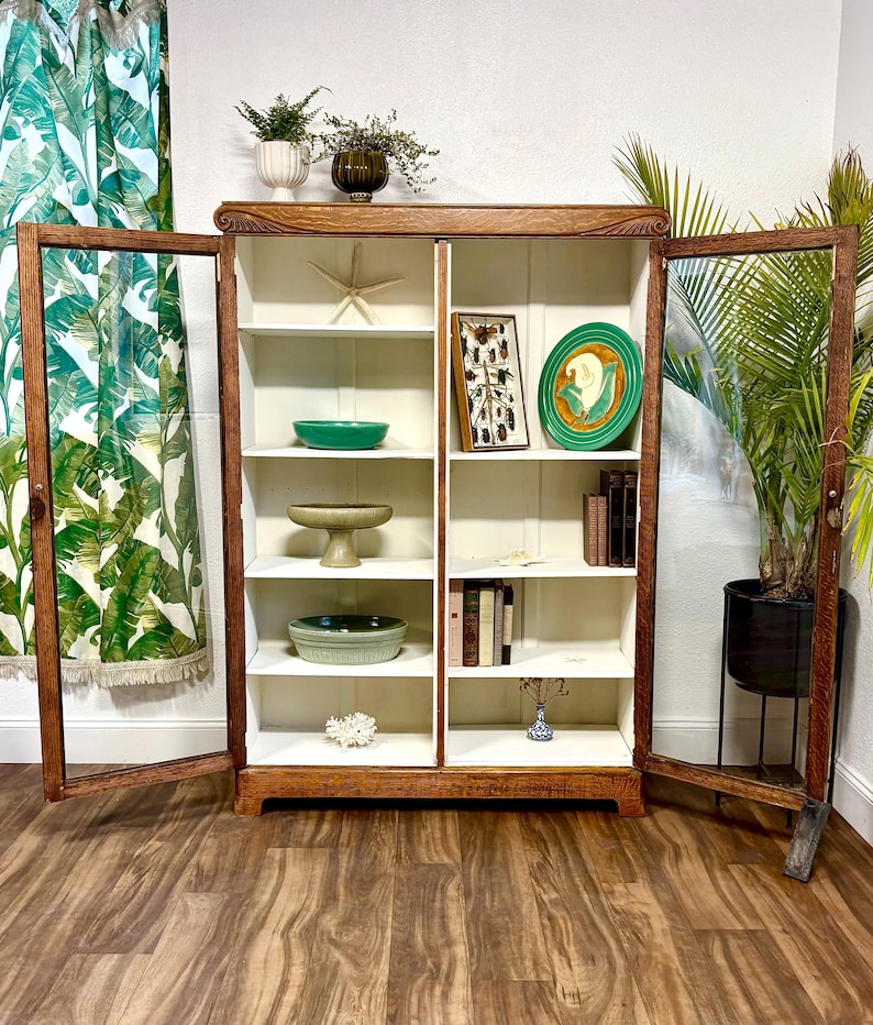 K&ouml;nnte beinhalten: Ein Vintage-Holzschrank mit Glast&uuml;ren, der verschiedene dekorative Gegenst&auml;nde pr&auml;sentiert. Der Schrank hat eine warme braune Oberfl&auml;che, wei&szlig;e Innenregale und enth&auml;lt einen Seestern, gr&uuml;ne Schalen, B&uuml;cher und eine gerahmte Insektenausstellung. Eine Topfpflanze ist sichtbar.