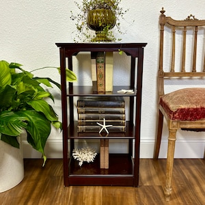 May include: A dark wood bookshelf with four shelves. The top shelf has a green ceramic planter with a leafy plant. The second shelf has a stack of books. The third shelf has a stack of books and a white starfish. The bottom shelf has a white coral and a stack of books.