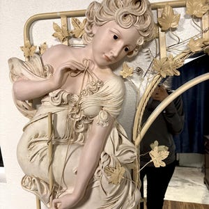May include: A white and cream colored decorative mirror with a sculpted woman figure. The woman is wearing a flowing dress and has a vine and leaf design around her and the mirrored portion.
