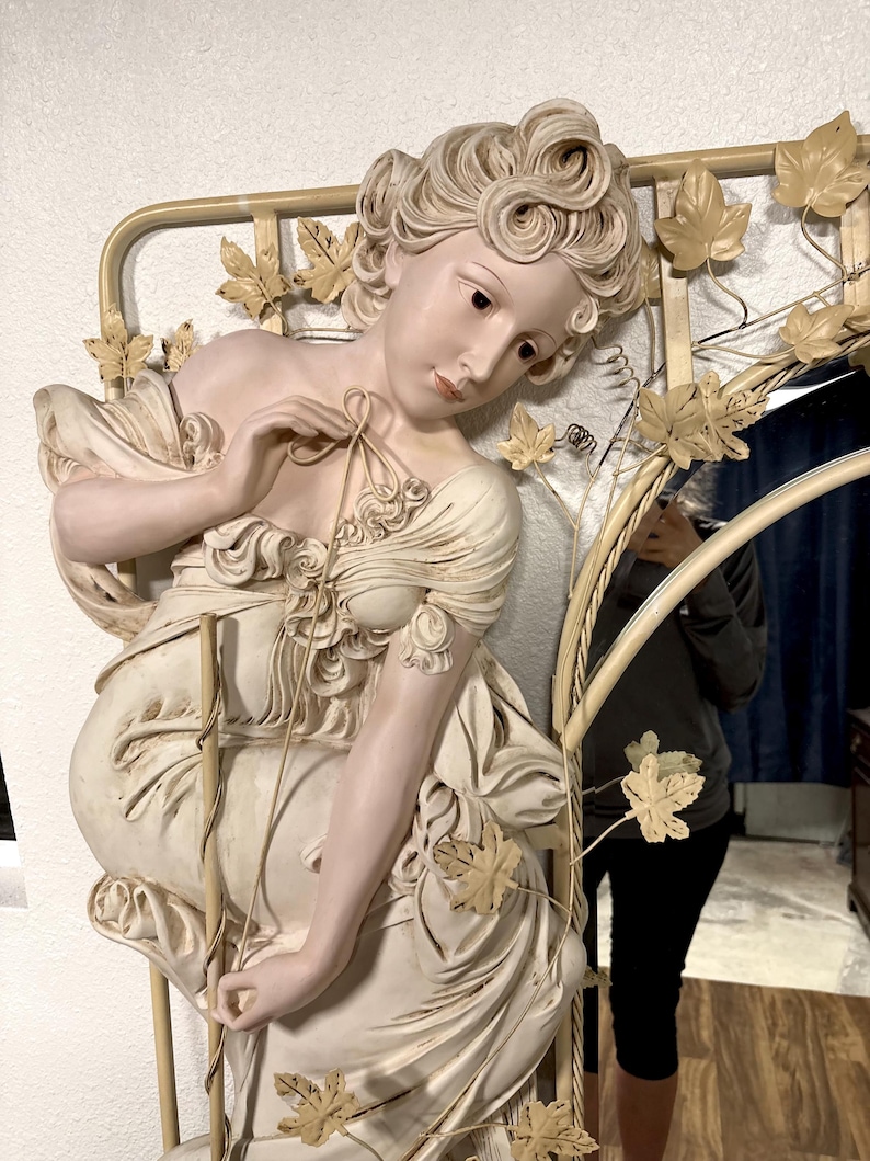 May include: A white and cream colored decorative mirror with a sculpted woman figure. The woman is wearing a flowing dress and has a vine and leaf design around her and the mirrored portion.