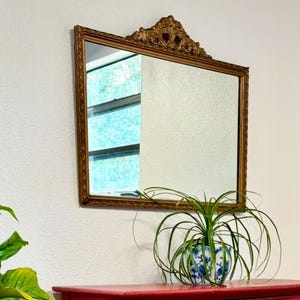 May include: A rectangular gold-framed mirror with ornate detailing at the top is mounted on a white wall. The mirror reflects a window. A red dresser with a blue and white patterned pot with a green plant sits below the mirror.