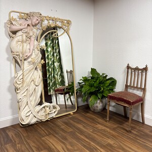 May include: A large, ornate mirror with a white, sculpted figure of a woman in a flowing gown. The mirror has a gold frame and is set against a white wall. A chair and a potted plant are in the background.