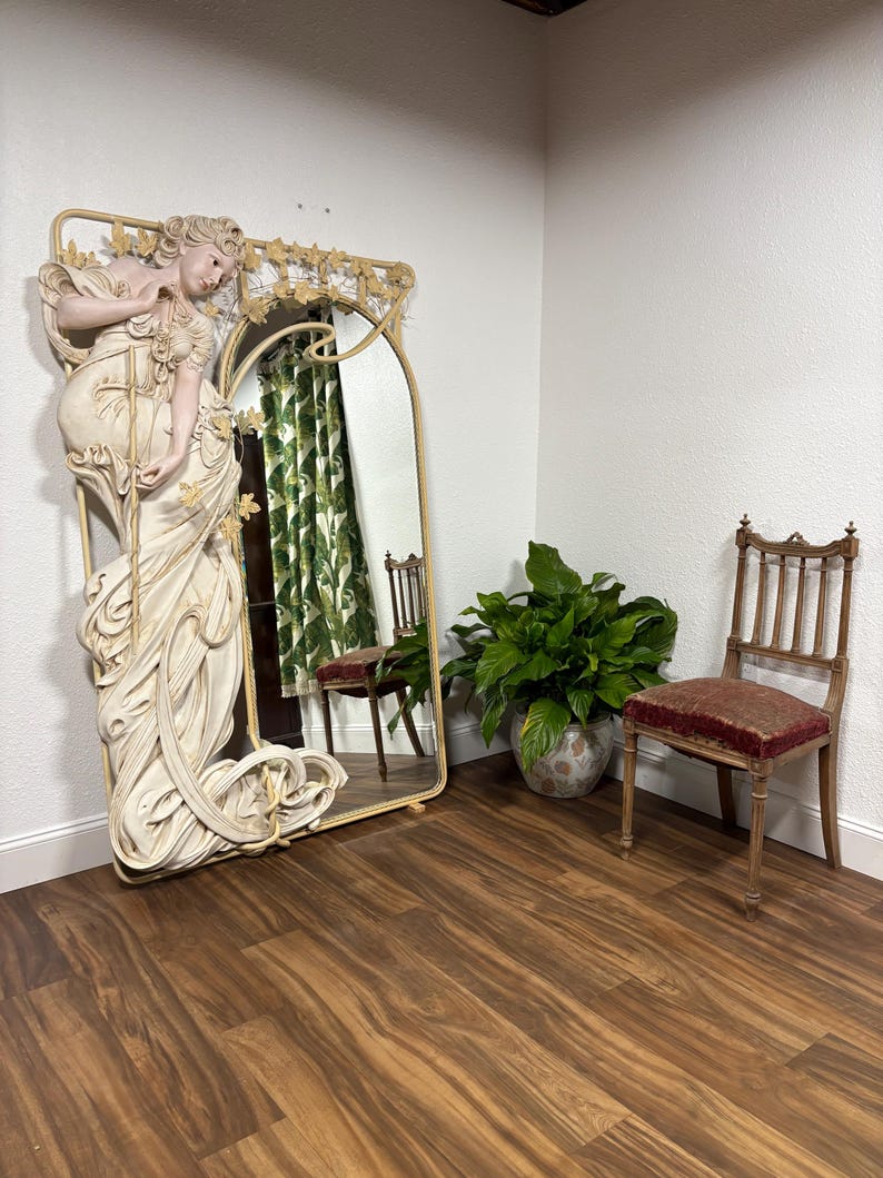 May include: A large, ornate mirror with a white, sculpted figure of a woman in a flowing gown. The mirror has a gold frame and is set against a white wall. A chair and a potted plant are in the background.