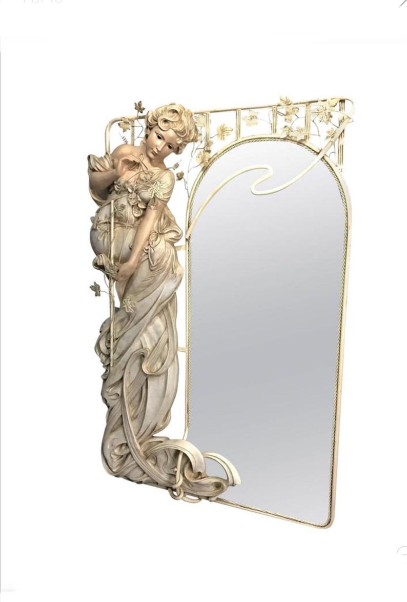 May include: A white, ornate mirror with a sculpted figure of a woman in a flowing gown. The mirror has a decorative frame with floral motifs and a curved top.