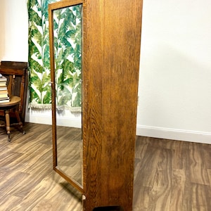 K&ouml;nnte beinhalten: Ein hoher Holzschrank mit Glast&uuml;r, der eine warme, mittelbraune Oberfl&auml;che aufweist. Die Schrankt&uuml;r ist ge&ouml;ffnet und gibt den Blick ins Innere frei. Die Holzmaserung ist sichtbar und verleiht Textur. Der Schrank steht vor einem neutralen Hintergrund.