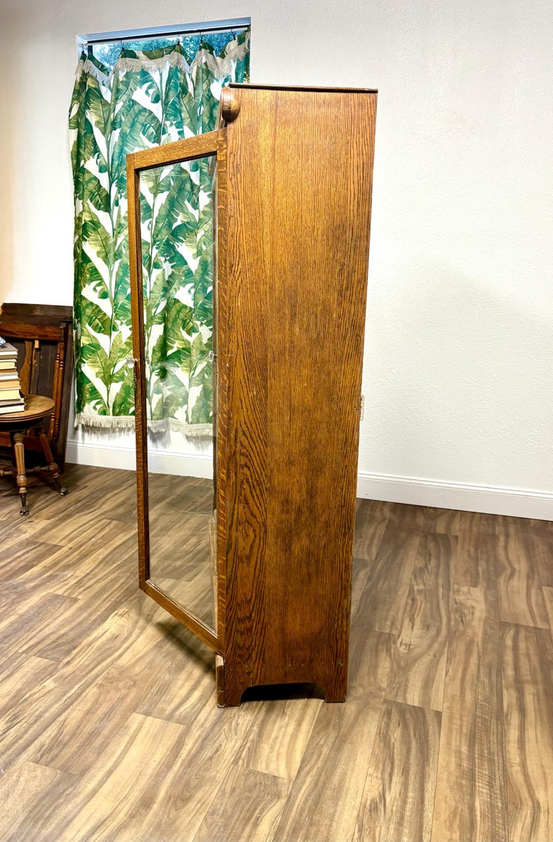 K&ouml;nnte beinhalten: Ein hoher Holzschrank mit Glast&uuml;r, der eine warme, mittelbraune Oberfl&auml;che aufweist. Die Schrankt&uuml;r ist ge&ouml;ffnet und gibt den Blick ins Innere frei. Die Holzmaserung ist sichtbar und verleiht Textur. Der Schrank steht vor einem neutralen Hintergrund.
