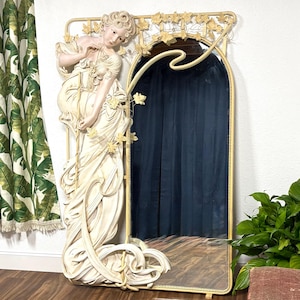 May include: A large, ornate mirror with a white, sculpted frame featuring a woman in a flowing gown. The frame is decorated with leaves and vines. The mirror has a gold-colored trim.
