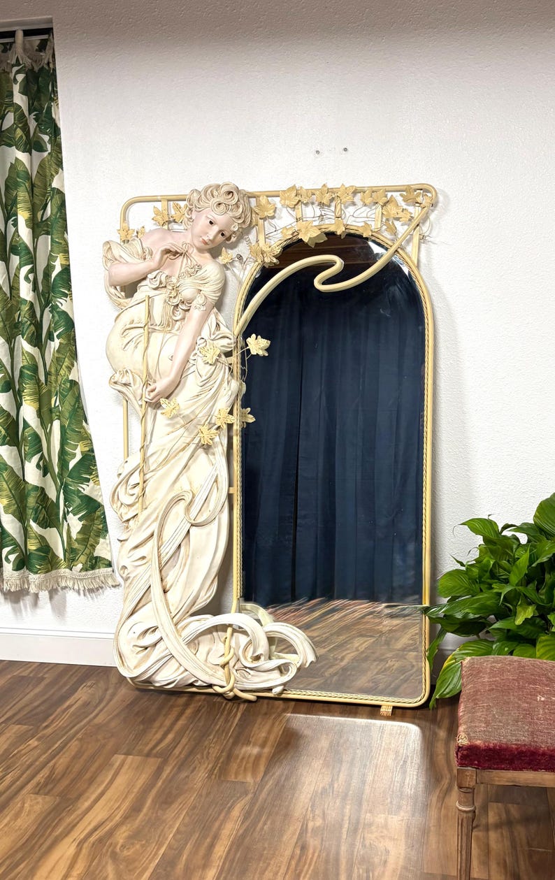 May include: A large, ornate mirror with a white, sculpted frame featuring a woman in a flowing gown. The frame is decorated with leaves and vines. The mirror has a gold-colored trim.