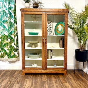 5’ tall Antique Art Nouveau Oak cabinet with carvings at the top corners. It has 2 glass doors and 8 shelves allowing for storage of large or small items. The natural warm brown Oak has beautiful wood grain and the interior is painted white. This versatile case piece could be used as a bookcase, food pantry, collectibles display, china cabinet, or even bathroom and / or linen storage. It is 59.5” Tall, 42.75” Wide, and 13.75” Deep.