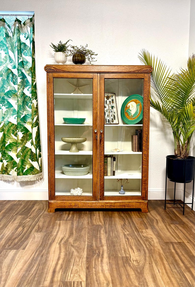5’ tall Antique Art Nouveau Oak cabinet with carvings at the top corners. It has 2 glass doors and 8 shelves allowing for storage of large or small items. The natural warm brown Oak has beautiful wood grain and the interior is painted white. This versatile case piece could be used as a bookcase, food pantry, collectibles display, china cabinet, or even bathroom and / or linen storage. It is 59.5” Tall, 42.75” Wide, and 13.75” Deep.