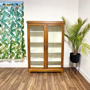K&ouml;nnte beinhalten: Ein hellbrauner Holzschrank mit Glast&uuml;ren und Regalen. Der Schrank hat eine dekorative Oberseite. Rechts befindet sich eine Topfpflanze in einem schwarzen Topf. Der Boden hat ein Holzmaserungsmuster. Ideal f&uuml;r Ausstellungsst&uuml;cke.