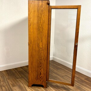 K&ouml;nnte beinhalten: Ein hoher Holzschrank mit einer halb ge&ouml;ffneten Glast&uuml;r. Der Schrank besteht aus hellem Braunholz mit einem ausgepr&auml;gten Maserungsmuster. Der T&uuml;rrahmen ist ebenfalls aus Holz und das Glas ist klar. Der Schrank steht vor einer wei&szlig;en Wand.