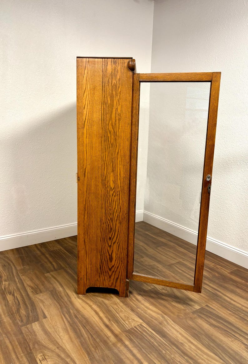 K&ouml;nnte beinhalten: Ein hoher Holzschrank mit einer halb ge&ouml;ffneten Glast&uuml;r. Der Schrank besteht aus hellem Braunholz mit einem ausgepr&auml;gten Maserungsmuster. Der T&uuml;rrahmen ist ebenfalls aus Holz und das Glas ist klar. Der Schrank steht vor einer wei&szlig;en Wand.