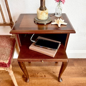 May include: A dark wood end table with a square top and a lower shelf featuring curved legs. On the top sits a brass and stone lamp, a small glass vase with pink flowers, and a white starfish. A smartphone rests on a book on the lower shelf.
