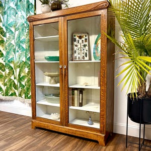 This is a large 5’ tall Antique American Oak cabinet with Art Nouveau style carvings at its’ shoulders. It has 2 glass doors and 8 adjustable  shelves allowing for storage of varying size items. The exterior is a natural warm brown Oak with beautiful wood grain and the interior is painted white.
This super versatile piece could be used as a bookcase, food pantry, display / china cabinet, or perhaps bath and linen storage.  59.5” Tall, 42.75” Wide, 13.75” Deep
