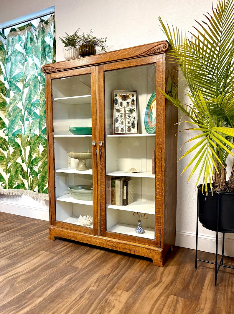 This is a large 5’ tall Antique American Oak cabinet with Art Nouveau style carvings at its’ shoulders. It has 2 glass doors and 8 adjustable  shelves allowing for storage of varying size items. The exterior is a natural warm brown Oak with beautiful wood grain and the interior is painted white.
This super versatile piece could be used as a bookcase, food pantry, display / china cabinet, or perhaps bath and linen storage.  59.5” Tall, 42.75” Wide, 13.75” Deep