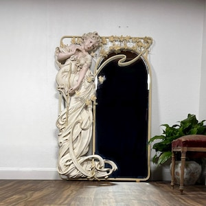 May include: A large, ornate mirror with a cream color iron frame and a life-size sculpted figure of a woman with flowing hair and a dress. The mirrors’ reflective surface has been blacked out and is surrounded by intricate floral details.