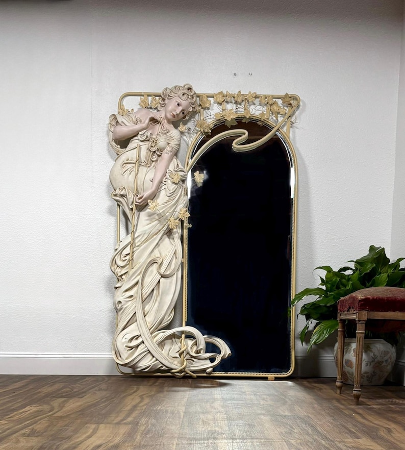 May include: A large, ornate mirror with a cream color iron frame and a life-size sculpted figure of a woman with flowing hair and a dress. The mirrors’ reflective surface has been blacked out and is surrounded by intricate floral details.