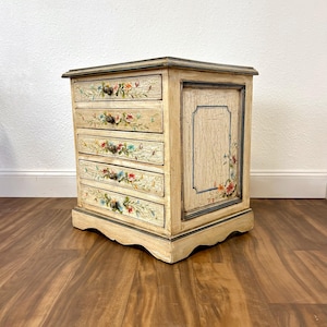 May include: A small, antique-style wooden chest with five drawers and a single door. The chest is painted in a light cream color and decorated with hand-painted floral designs in shades of pink, blue, and green.